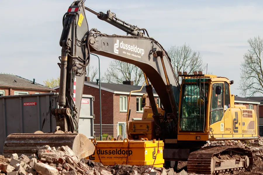 Gele graafmachine op een bouwplaats bij Nederlandse rijtjeshuizen, voorbeeld van graafwerk waarvoor een KLIC-melding nodig is